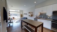 This single-floor space showcases a contemporary open-concept layout that seamlessly integrates the kitchen, dining, and living areas into one cohesive environment. The kitchen is appointed with white cabinetry, a gray subway tile backsplash, stainless steel appliances, and a sink positioned along the countertop. Recessed lighting illuminates the space from above, complementing the bright, airy atmosphere created by large floor-to-ceiling windows that allow generous natural light to flood in from one end of the room. The dining area features a substantial dark wood table with a lower storage shelf, paired with modern seating. The living area includes a casual seating arrangement set atop a neutral area rug over warm hardwood floors. Wall-mounted open shelving provides functional display and storage space. The overall design aesthetic is minimalist and modern, characterized by a crisp white and gray palette, clean architectural lines, and a well-organized layout that maximizes the sense of openness throughout the space.
