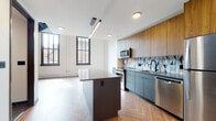 Located on Euclid Avenue in the heart of Cleveland, Ohio, this two-floor residence blends contemporary design with thoughtful architectural detail throughout. The main level showcases a striking open-concept kitchen and living area, anchored by herringbone-patterned wood flooring that adds warmth and visual interest. The kitchen is a standout feature, outfitted with warm wood upper cabinetry, matte gray lower cabinets, stainless steel appliances including a refrigerator, range, microwave, and dishwasher, and a bold geometric-patterned tile backsplash. A large central island with a sleek gray countertop provides additional workspace and serves as a focal point for the room. Tall, black-framed windows flood the living area with natural light and frame views of the surrounding urban landscape. A modern staircase with a black metal railing leads to the second floor, which offers an open loft-style space with wood flooring and recessed lighting. The bathrooms are finished with hexagonal tile flooring, white subway tile shower surrounds, wood-toned vanities, white countertops, and matte black fixtures throughout, creating a cohesive and contemporary aesthetic. The overall design reflects a refined industrial sensibility, combining clean lines, mixed materials, and modern finishes.