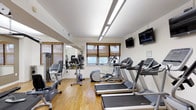 This fitness center is a thoughtfully designed, single-floor exercise space built to support a well-rounded workout routine. The room features warm hardwood flooring throughout, which adds a clean and inviting aesthetic to the functional environment. Natural light filters in through multiple windows dressed with woven bamboo shades, complemented by overhead fluorescent lighting that ensures the space remains bright and well-lit at all times. A large wall mirror on one side visually expands the room and allows users to monitor their form during exercise. The cardio equipment lineup includes multiple treadmills, an elliptical trainer, and stationary bikes, offering a variety of options for aerobic workouts. A dedicated free weights area is visible along the mirrored wall, providing options for strength training. Two wall-mounted flat-screen televisions are positioned for easy viewing from the cardio machines, enhancing the workout experience. The layout is open and efficiently organized, allowing for comfortable movement between equipment. The overall ambiance is clean, functional, and welcoming, making it a practical and well-appointed space for daily fitness use.