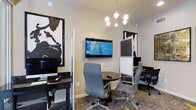 This single-floor business center offers a thoughtfully designed workspace that balances functionality with modern aesthetics. The space features a clean, contemporary interior with neutral walls that serve as a backdrop for bold black-and-white abstract artwork, adding a striking visual element to the professional environment.

The layout is intelligently divided to accommodate both individual and collaborative work styles. Dedicated computer workstations with desktop monitors are positioned along the perimeter, providing focused areas for productivity. At the center of the room, a round black table surrounded by ergonomic gray office chairs with polished chrome bases creates an inviting hub for small meetings or group work sessions. A wall-mounted flat-screen television serves as a presentation or information display, enhancing the room's utility.

Privacy partitions and dividers help section off distinct work zones, offering users a degree of separation without sacrificing the open feel of the space. Overhead, a modern sputnik-style chandelier with globe bulbs provides warm, ambient lighting that complements the room's professional yet approachable atmosphere. Patterned carpet tiles complete the interior, adding texture and visual interest underfoot. Overall, this business center is well-suited for focused work, small meetings, and collaborative sessions alike.