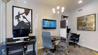 This single-floor business center offers a thoughtfully designed workspace that balances functionality with modern aesthetics. The space features a clean, contemporary interior with neutral walls that serve as a backdrop for bold black-and-white abstract artwork, adding a striking visual element to the professional environment.

The layout is intelligently divided to accommodate both individual and collaborative work styles. Dedicated computer workstations with desktop monitors are positioned along the perimeter, providing focused areas for productivity. At the center of the room, a round black table surrounded by ergonomic gray office chairs with polished chrome bases creates an inviting hub for small meetings or group work sessions. A wall-mounted flat-screen television serves as a presentation or information display, enhancing the room's utility.

Privacy partitions and dividers help section off distinct work zones, offering users a degree of separation without sacrificing the open feel of the space. Overhead, a modern sputnik-style chandelier with globe bulbs provides warm, ambient lighting that complements the room's professional yet approachable atmosphere. Patterned carpet tiles complete the interior, adding texture and visual interest underfoot. Overall, this business center is well-suited for focused work, small meetings, and collaborative sessions alike.