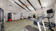 Located in Aurora, Colorado, this fitness center offers a well-appointed exercise space designed to support a variety of workout routines. The room features soaring vaulted ceilings accented by bold dark wood beams, creating an open and energizing atmosphere. Stylish pendant light fixtures provide warm, even illumination throughout the space, while a large multi-panel window wall floods the room with natural light and offers views of the surrounding exterior.

The floor is covered in a contemporary modular carpet tile in a mix of gray and yellow-green tones, adding visual interest underfoot. A full-length mirror along one wall enhances the sense of depth and allows users to monitor their form during workouts.

Equipment includes multiple treadmills, an elliptical trainer, a stationary cycle, a cable machine, a dumbbell rack with a full range of free weights, and additional strength and functional training accessories. A wall-mounted television is also present for entertainment during cardio sessions.

Adding a distinctive local character, the walls are decorated with logos and graphics representing Colorado's professional sports teams, including the Avalanche, Broncos, Rockies, and Rapids, giving the space a spirited, community-oriented feel. A frosted glass partition near the entrance provides a sleek architectural detail. This fitness center is a functional and inviting space for residents to maintain an active lifestyle.