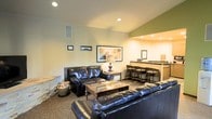 This single-floor space offers a well-appointed and inviting layout designed for comfortable living and entertaining. The main living area is anchored by a pair of plush black leather sofas arranged around a glass-top coffee table, creating an ideal gathering spot. Vaulted ceilings add a sense of openness and volume to the room, while recessed lighting provides warm, even illumination throughout. The walls are finished in neutral tones accented by a rich green feature wall that adds a pop of color and visual interest. Framed artwork decorates the walls, contributing to the space's polished, lived-in character. A stone-accented architectural element along one wall adds a rustic, textural touch. Adjacent to the living area, a built-in wet bar or kitchenette features bar-height seating with wicker-back stools, a countertop workspace, and overhead recessed lighting, making it a practical and stylish spot for casual dining or entertaining. The carpeted flooring throughout adds warmth and comfort underfoot. Overall, the space strikes a balance between relaxed comfort and functional design, making it well-suited for both everyday use and hosting guests.