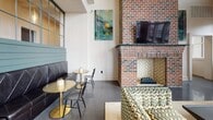 Located at Livingston Apartment Flats in Chesterfield, Virginia, this single-floor clubhouse is a thoughtfully designed community gathering space that blends industrial character with refined comfort. The main lounge area commands attention with soaring double-height ceilings and a dramatic full-height brick fireplace accented by a herringbone tile surround and a mounted flat-screen television above the mantle. Chevron-patterned accent chairs and curated artwork contribute to the space's eclectic, design-forward personality. A shuffleboard table and pool table add recreational appeal to the lounge area, complemented by cozy banquette seating with gold bistro tables. The kitchen features dark tile-clad island counters, sage green cabinetry with dark hardware, and polished concrete floors, with direct access to an outdoor patio. Floor-to-ceiling windows along one wall flood the space with natural light and offer open views of the surrounding community. A wide corridor with warm herringbone hardwood flooring, vaulted ceilings, a sculptural chandelier, and a row of upholstered chairs with bold orange accents connects the gathering areas. Throughout, polished concrete floors, curated art, and distinctive branded signage reinforce a cohesive, vibrant aesthetic ideal for resident socializing and events.