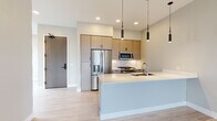 This contemporary single-floor residence showcases a clean, modern design with a thoughtful open-concept layout. The living and kitchen areas flow seamlessly together, anchored by light wood-tone flooring that lends warmth throughout. The kitchen is a standout feature, outfitted with flat-front cabinetry in a natural wood finish, white subway tile backsplash, stainless steel appliances including a French door refrigerator, a gas range, and a built-in microwave. A generous peninsula with a crisp white countertop provides both prep space and casual seating, complemented by sleek pendant lighting overhead. The open living area is bright and airy, benefiting from large sliding glass doors and adjacent windows that invite ample natural light and offer views of the surrounding neighborhood. The carpeted bedroom continues the light, neutral palette and features a ceiling fan and large windows that further enhance the sense of openness. The full bathroom is finished with marble-look tile flooring, a white subway-tiled tub and shower combination, and modern chrome fixtures. A spacious walk-in closet with built-in shelving and hanging rods provides generous storage. Recessed lighting throughout ties the design together with a polished, cohesive aesthetic.