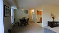 This single-floor residence offers a practical, open layout that connects key living areas seamlessly. The dining area is a notable focal point, centered around a round, dark-finished table accompanied by four matching chairs. A pendant light hangs directly above, providing focused illumination, while a framed piece of wall art adds a decorative touch. Neutral-toned carpeting spans the main living and dining areas, contributing to a warm and cohesive atmosphere throughout.

Adjacent to the dining space, the kitchen is partially open to the main area and features warm, medium-toned wood cabinetry on both upper and lower sections. A stainless steel dishwasher and a single-basin sink with a standard faucet are visible, alongside additional lower cabinetry that provides ample storage. The kitchen's layout suggests efficient use of space without sacrificing functionality.

A central hallway extends from the dining area toward what appears to be additional rooms, offering a clear sense of depth and flow within the home. The muted, neutral wall tones throughout complement the carpeted floors and create a calm, understated ambiance suited to everyday living.