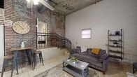 Located at 601 Bridge St in Danville, Virginia, this two-story loft blends historic industrial character with modern functionality. The space is defined by its striking exposed brick walls, raw concrete and vaulted ceilings, and light-toned tile flooring that carry throughout both levels, creating a cohesive urban aesthetic.

The main floor houses an open living room anchored by tall ceilings and large vertical windows that draw in natural light against a whitewashed brick backdrop. A galley-style kitchen features white cabinetry, light countertops, stainless steel appliances, and a classic brick accent wall. A dedicated laundry room and two closets add practical storage to the layout.

A black metal staircase connects the two floors, lending an industrial flair to the transition. The expansive upper-level bedroom offers generous proportions, a ceiling fan, arched brick doorways, and vaulted ceilings that amplify the sense of volume. The bathroom continues the brick motif with exposed masonry walls paired with clean tile flooring.

Two closets provide ample storage, while the overall layout—spanning two distinct floors—offers a sense of separation between living and sleeping areas rarely found in similarly sized spaces. The building's historic bones and thoughtful conversion make this a distinctive urban retreat.