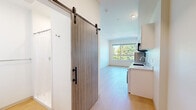 Located at 4612 Stone Way N in Seattle's vibrant Wallingford neighborhood, this modern studio is thoughtfully designed to maximize every inch of its compact footprint. The open-concept layout seamlessly connects the living area and kitchen, creating an efficient and comfortable living environment. Light hardwood flooring runs throughout, complementing the crisp white walls and recessed lighting that keep the space feeling bright and airy.

The well-appointed kitchen lines one wall with white high-gloss cabinetry extending to the ceiling for ample storage. It is fully equipped with stainless steel appliances including a refrigerator, microwave, electric cooktop, dishwasher, and a conveniently integrated in-unit washer — all finished with a clean white countertop and stainless steel sink.

A stylish sliding barn door with dark hardware separates the bathroom from the main living area, adding a modern architectural touch. The bathroom features light hardwood flooring, a clean white palette, a shower/tub combination, and brushed nickel fixtures.

The living area is anchored by a large picture window that frames lush, green tree canopy views, flooding the room with natural light and bringing a sense of the outdoors inside. A bold red entry door adds a striking accent upon arrival. This well-designed studio offers a clean, contemporary aesthetic ideal for efficient urban living in Seattle.