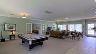 This well-appointed single-floor space is designed for both recreation and relaxation, offering a generous open layout that combines a game area with a comfortable lounge. The room is anchored by a full-sized pool table positioned on light-toned tile flooring, making it a natural focal point for entertainment and social gatherings.

Transitioning seamlessly from the game area, a carpeted lounge zone features wicker and upholstered seating arranged around a television setup, creating an inviting space for unwinding. Lush indoor plants are thoughtfully placed throughout, adding a refreshing, natural element to the interior.

Large windows along the far wall flood the space with natural light, enhancing the airy, open ambiance. The soft sage-green walls paired with white crown molding and a textured ceiling contribute to a calm and welcoming atmosphere. Pendant light fixtures provide warm supplemental lighting throughout.

French doors with glass panes connect this main area to an adjoining room, offering both visual continuity and functional separation. The combination of tile and carpet flooring zones the space naturally, while the overall design balances casual comfort with recreational functionality — making it an ideal communal or leisure space.