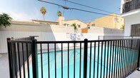 This single-floor unit is part of a well-maintained multi-unit residential building with a distinctly Southern California aesthetic. The building's exterior is finished in crisp white stucco, lending a clean, modern look that is accentuated by ornamental black wrought iron details throughout — visible on balconies, stair railings, window security guards, and entry gates. The unit's entrance is accessed via a short flight of grey-painted concrete steps, flanked by a decorative iron handrail, and secured by a matching ornate iron security door, conveying both style and safety.

The building is oriented around a central courtyard that houses a generously sized shared swimming pool with clear turquoise water, enclosed by a black iron safety fence. The pool area is surrounded by a tiled deck and is open to the sky, allowing abundant natural sunlight throughout the day. Palm trees are visible above the perimeter walls, reinforcing the warm, sun-drenched Southern California atmosphere.

Exterior walkways and corridors are finished in smooth concrete, providing clean circulation paths between units. Upper-level balconies with decorative iron railings overlook the pool courtyard, adding architectural dimension and visual interest. Overall, the property presents a cohesive, resort-inspired ambiance well-suited to warm-weather living.