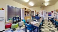 Located at 3131 Simpson Stuart Rd in Dallas, TX, this single-floor business center and activity room is a well-equipped, multifunctional space designed to support both productivity and community engagement. The room features a striking vaulted ceiling that adds a sense of openness and volume, complemented by bright fluorescent lighting throughout. A bold black-and-white checkered tile floor runs the length of the space, giving it a lively, distinctive character.

The layout is organized into several functional zones. A dedicated business center area lines one wall with individual computer workstations, each equipped with a monitor and separated by dividers for focused use, alongside a posted "Business Center Etiquette" sign. Large communal dark-wood tables with teal and blue upholstered chairs occupy the central floor area, providing ample space for group activities, meetings, or study sessions.

A bay window nook at the far end of the room offers natural light through tall windows fitted with wood-toned blinds, creating a cozy alcove with a small table and chairs. Built-in and freestanding shelving units along the walls provide extensive storage for books, supplies, and resources. A colorful reading and lounge corner features modular ottomans in green and white. Bulletin boards, wall décor, and seasonal displays throughout contribute to the room's warm, welcoming, and community-oriented atmosphere.