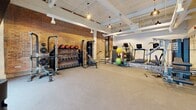 Located at 450 Amsterdam St in Detroit, Michigan, this fitness center combines industrial character with modern functionality. The space is defined by its exposed brick walls, white-painted structural beams, and an open ceiling with visible ductwork, all contributing to a loft-inspired aesthetic. Pendant lighting fixtures are suspended throughout, casting warm, focused light across the workout floor.

The gym is generously laid out across a single level, providing ample room for a wide variety of fitness activities. The cardio section features multiple treadmills and elliptical machines, each equipped with individual screens for an interactive workout experience. A dedicated cycling area includes Echelon spin bikes for high-intensity training sessions. Strength training is well represented with a multi-station cable machine, weight racks stocked with kettlebells and medicine balls, barbells, and a selection of benches.

Large, grid-paned windows along the exterior walls allow natural light to fill the space and offer views to the outdoors, enhancing the open, airy feel. Full-length mirrors on select walls visually expand the room and support proper form during workouts. Stability balls and functional training accessories round out the equipment offerings. The neutral-toned flooring provides a clean, cohesive foundation throughout. The overall ambiance is energetic yet inviting, making it a comprehensive facility for a wide range of fitness needs.