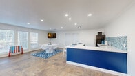 This 630 square foot single-floor living space in Tomball, TX, offers a blend of modern design and functionality. The open-concept layout maximizes the use of space, creating a seamless flow between different areas. Large windows with white blinds allow abundant natural light to flood the room, enhancing the sense of spaciousness.

The flooring throughout is a striking earth-toned tile, adding warmth and durability to the space. A standout feature is the custom-built blue island with a white countertop, which serves as both a kitchen workspace and a casual dining area, complete with modern white bar stools.

The kitchen area boasts a mosaic tile backsplash in shades of blue, complementing the island and adding a touch of visual interest. Recessed lighting fixtures are strategically placed throughout the ceiling, ensuring even illumination across the space.

In one corner, a unique accent wall covered in artificial greenery with the word "Budget" displayed adds a pop of nature and personality to the room. The living area is versatile, with ample space for seating and entertainment, including a wall-mounted TV.

The room's neutral white walls provide a blank canvas for personalization, while blue accents in the form of a geometric rug and furniture pieces tie the color scheme together. This thoughtfully designed space strikes a balance between style and practicality, suitable for various lifestyle needs.