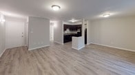 This single-floor unit is designed with a clean, functional layout that connects the main living spaces in a practical and cohesive manner. The open-concept living area is finished with light wood-look luxury vinyl flooring, which extends throughout the main areas and bathroom, lending a bright and cohesive feel to the space. Large sliding glass doors and a wide window allow natural light to fill the living area while offering views of the surrounding outdoor greenery.

The kitchen is equipped with dark espresso cabinetry, granite-style countertops, and black appliances, including a refrigerator, range, and dishwasher. A peninsula provides additional counter space and subtly defines the kitchen from the adjacent living area. An under-cabinet range hood and a stainless steel sink complete the functional setup.

The bedroom features neutral carpet flooring, soft grey walls, and a standard closet with a hanging rod. A window offers additional natural light and outdoor views. The full bathroom includes a tub-shower combination with white tile surround, a toilet, and a dark vanity, all set on the same light wood-look flooring found throughout the unit. The overall aesthetic is modern and understated, with a neutral palette that creates a versatile and welcoming atmosphere.