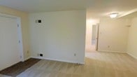 This single-floor space presents a straightforward and functional layout finished in neutral tones throughout. The main living area features wall-to-wall carpeting in a light beige tone, contributing to a warm and understated atmosphere. The walls are painted in a crisp off-white, providing a clean, blank canvas. A partial wall divides the space, creating a subtle sense of separation between areas while maintaining an open feel. Ventilation registers are positioned both on the wall and near the baseboard, indicating a dual HVAC system for efficient climate control. A standard electrical outlet is visible along the baseboard, offering convenient access. Beyond the partial dividing wall, a hallway leads to additional rooms, which appear to continue the same neutral carpet and paint scheme. The connected room visible in the background is well-lit, suggesting either a window or overhead lighting source nearby. White-painted door frames and baseboards provide clean visual definition throughout. Overall, the space offers a simple, move-in-ready environment with consistent finishes and a practical, accessible layout across its single floor.