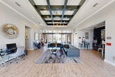 Located at 24215 Kuykendahl Road in Tomball, Texas, this sophisticated single-floor space makes an immediate impression with its striking coffered ceiling — a bold geometric grid finished in dark tones and accented with recessed lighting and an elegant chandelier. The open-concept layout allows the space to flow seamlessly from one zone to the next, all unified by warm-toned hardwood-style flooring and a crisp, neutral color palette.

The central lounge area is anchored by a pair of tufted leather sofas arranged around a glass-top coffee table atop a large abstract area rug, creating an inviting and stylish gathering space. Large floor-to-ceiling windows and glass-paneled entry doors along the front facade flood the interior with abundant natural light while offering clear views to the outdoors.

To one side, a dedicated bar area with high-top seating and dark accent walls adds a social, upscale ambiance. Framed artwork, a sunburst mirror, and decorative accessories are thoughtfully placed throughout, contributing to the refined aesthetic. A secondary alcove with additional seating and a workstation provides a functional counterpoint to the lounge.

The overall atmosphere is modern and polished, blending contemporary design elements with warm, welcoming touches — making this a well-appointed space ideal for community gatherings or social events.