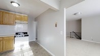 This single-floor residence presents a straightforward and functional layout designed for comfortable everyday living. The kitchen is equipped with a gas range and features light wood cabinetry, offering ample storage and a practical workspace. Neutral-toned walls and light-colored tile flooring run throughout the space, creating a bright and cohesive aesthetic that is easy to personalize. The open transition between the kitchen and adjoining living area allows for a natural flow, making the space feel connected and accessible. A decorative wall sconce adds a subtle design touch to the hallway area, while clean white baseboards and trim provide a crisp finish throughout. The living area is bathed in ambient light and features an ornamental wrought iron stair railing as a visual accent, adding a classic architectural detail to the interior. The overall layout is efficient and well-organized, with clear sightlines between rooms that enhance the sense of openness. The neutral palette and clean finishes make this a versatile space, well-suited for a variety of interior styles and furnishing preferences.