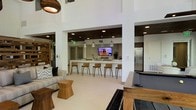 This single-floor space is designed as a multifunctional communal area with a clean, contemporary aesthetic. The interior is characterized by soaring ceilings and crisp white walls that create an open, airy atmosphere throughout. Recessed lighting and pendant fixtures provide layered illumination across the space.

A central kitchen bar serves as a focal point, featuring a sleek white countertop with wood-backed bar stools and pendant lighting overhead. The bar area is equipped with a wall-mounted flat-screen television, making it ideal for social gatherings. Adjacent to the bar, a stainless steel refrigerator and additional cabinetry offer practical functionality. The ceiling above the bar area features a warm wood finish, adding contrast to the otherwise bright palette.

The lounge area is furnished with a neutral-toned sofa accented with patterned throw pillows, complemented by a geometric wood side table. A game area is also present, featuring a pool table and area rug, adding an entertainment dimension to the space. A bookshelf visible in the background further enhances the space's versatile, social character. Overall, the design balances modern minimalism with warm wood accents.