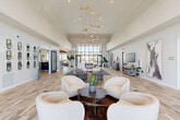 Located at 28851 N Vistancia Blvd in Peoria, Arizona, this single-floor community clubhouse offers a generously sized, thoughtfully designed gathering space. The centerpiece of the main living area is a dramatic cascading gold chandelier that draws the eye upward toward soaring ceilings, filling the room with a sense of grandeur. Large floor-to-ceiling windows at the entrance flood the interior with natural light and provide views of the surrounding Arizona landscape.

Light-toned wood-look tile flooring flows seamlessly throughout, unifying the open-concept layout. Multiple distinct seating areas are arranged across the expansive main floor, including plush curved lounge chairs, modular sofas, and a reception-style desk, making the space equally suited for socializing, working, or relaxing. Curated artwork and lush indoor plants add warmth and visual interest throughout.

A fully appointed kitchenette area features a center island with bar seating, pendant lighting, stainless steel appliances, and ample counter space, ideal for community gatherings. A dedicated media lounge, finished in deep teal tones with tray ceiling accent lighting, provides an intimate setting with multiple mounted televisions and comfortable seating. A welcoming entryway corridor with a beverage station and decorative vignettes completes this well-appointed community space.