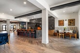 This expansive single-story clubhouse spans a generous footprint designed to serve as a dynamic community gathering space. The entrance lobby makes an immediate impression with its warm wood-paneled ceiling, bold exposed beams, and large-format tile flooring. A hand-painted mountain mural anchors one wall, while a reception desk and lounge seating create a welcoming arrival experience.

Moving deeper into the clubhouse, the layout opens into a spacious social lounge featuring herringbone wood-look tile flooring, dark exposed ceiling beams, and a dramatic floor-to-ceiling stone fireplace with an integrated television. Generous sectional seating, layered rugs, and curated furnishings establish a comfortable yet refined atmosphere.

Adjacent to the lounge, a full-length bar area commands attention with its teal cabinetry, dark stone countertops, and a striking wall of vertical metallic tile. Bar stools line a peninsula counter, creating an inviting setting for casual socializing. A pool table nearby adds a recreational dimension to the space.

Cozy dining nooks with booth-style seating and pendant lighting offer more intimate settings, while floor-to-ceiling windows throughout flood the interior with natural light and provide views of the surrounding landscape. The overall design balances a modern aesthetic with warm, lodge-inspired materials.