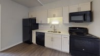 This single-floor unit offers a practical and well-organized layout, as seen in the 3D floor plan, which reveals a seamless flow between the main living spaces. The open-concept area connects the kitchen and living room, creating a spacious and functional environment. Light-toned walls and hardwood-style flooring run throughout, contributing to a clean and cohesive aesthetic.

The kitchen is outfitted with white shaker-style cabinetry extending to the upper walls, providing ample storage. Black appliances — including a refrigerator, electric range, over-the-range microwave, and dishwasher — contrast neatly against the white cabinetry and light countertops. A double-basin stainless steel sink with a modern faucet adds a practical touch. Under-cabinet lighting illuminates the workspace effectively.

The living area is bright and open, benefiting from multiple windows fitted with security bars that allow natural light to filter in. A ceiling fan provides ventilation and additional lighting. A baseboard heating unit runs along the exterior wall.

The bathroom features floor-to-ceiling beige tile work, a soaking tub with an overhead shower, a decorative mosaic tile border, and a frosted window for natural light and privacy.