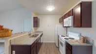 Located at 2332 Asics Dr in Hilliard, Ohio, this single-floor unit offers a practical and well-appointed layout designed for comfortable everyday living. The galley-style kitchen is a standout feature, showcasing rich dark cabinetry paired with granite-look countertops, a double basin sink, and white appliances including an electric range, refrigerator, and over-the-range microwave. Wood-look flooring runs throughout the kitchen, adding warmth and easy maintenance. A half-wall peninsula connects the kitchen to the main living area, creating an open, connected feel while maintaining a defined sense of space. The living area features neutral carpet, warm-toned walls, and a full sliding glass door dressed with vertical blinds, allowing natural light to filter in while providing access to outdoor views. A bedroom features plush carpeting, a window with blinds for natural light, and a textured ceiling, offering a quiet and functional space. The bathroom is cleanly finished with a full tub-shower combination, wood-look flooring, a granite-topped vanity with dark cabinetry, and brushed nickel fixtures. Throughout the unit, the consistent use of neutral tones and complementary finishes creates a cohesive and inviting atmosphere.