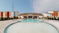 Located at 300 Bellamy Ave in Conway, South Carolina, this single-story community facility offers a well-appointed gathering space designed for relaxation and recreation. The centerpiece of the property is a large, sparkling outdoor swimming pool with a clean, white-tiled interior that reflects the open South Carolina sky. Surrounded by a generous pool deck, the area provides ample space for lounge seating, making it ideal for sunbathing and socializing. The clubhouse building features a classic design with a pitched, dark-shingled roof and crisp white exterior, anchored by a covered rear porch supported by columns. A series of glass-paned French doors lines the back facade, opening directly onto the patio and allowing natural light to flow into the interior while providing seamless indoor-outdoor connectivity. The covered patio is furnished with additional seating areas, creating a shaded retreat for residents. Red accent details on the doors and trim add a subtle pop of color to the otherwise neutral palette. The overall ambiance is clean, bright, and inviting, reflecting a well-maintained community amenity space suited for year-round enjoyment in the coastal South Carolina climate.