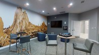 This single-floor lounge space is thoughtfully designed to create a comfortable and visually engaging environment for socializing and entertainment. The room's most striking feature is a large, floor-to-ceiling photographic mural depicting a dramatic rocky landscape, rendered in warm earth tones that contrast boldly against the cool slate-gray accent wall. Mounted on the accent wall is a flat-screen television, complemented by built-in media equipment and wall-mounted speakers, making the space well-suited for viewing and entertainment. Recessed ceiling lighting provides even, modern illumination throughout. The seating arrangement is varied and functional, featuring curved navy blue accent chairs with subtle LED lighting accents, sleek gray side chairs, and tall bar-height stools paired with round high-top tables. The neutral, low-pile carpeting grounds the space and contributes to its contemporary aesthetic. A white door in the corner offers access to adjacent areas. Overall, the space strikes a balance between a professional and casual atmosphere, with curated décor and versatile furnishings that make it suitable for both informal gatherings and structured activities.