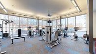 Located at 3190 Brighton Blvd in Denver, CO, this fitness center offers a thoughtfully designed exercise space with a generous single-floor layout. Floor-to-ceiling wraparound windows dominate the perimeter, flooding the room with natural light and providing open views of the surrounding Denver cityscape, creating an energizing atmosphere for workouts.

The space is comprehensively equipped to support a wide range of fitness routines. The cardio zone features multiple treadmills, elliptical trainers, stationary bikes, and a cable machine with an integrated display screen, all positioned to take advantage of the expansive street-level views. A ceiling fan overhead aids in ventilation and comfort during intense sessions.

The strength training area is equally well-appointed, housing a full rack of dumbbells, barbells with weight plates, a squat/power rack, a flat weight bench, and a dedicated functional training station complete with battle ropes, kettlebells, medicine balls, resistance bands, and TRX suspension equipment. A wall-mounted mirror enhances the sense of space in this zone.

A vibrant athletic mural depicting an outdoor mountain biking scene adds a dynamic, Colorado-inspired aesthetic to one wall, complementing the modern, urban industrial design of the facility. Patterned carpet tiles in neutral tones accented with blue geometric designs cover the floors throughout, tying together the clean, contemporary look of the space.