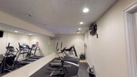Located at 5461 Collins Mill Creek Drive in Milton, Florida, this single-floor fitness center is designed to provide a functional and well-equipped workout environment. The space features a selection of cardio machines, including ellipticals and a treadmill, arranged to maximize usability within the room. A large wall-mounted mirror along one side creates a sense of openness and allows users to monitor their form during exercise. Recessed lighting in the textured ceiling provides bright, even illumination throughout the space. A wall-mounted flat-screen television adds an entertainment element, making workouts more engaging. The flooring consists of durable, dark rubber matting suited for high-traffic fitness use. A wall-mounted fan ensures air circulation and comfort during activity. A small decorative plant near the equipment adds a touch of warmth to the otherwise utilitarian setting. The neutral-toned walls and clean layout contribute to a focused, distraction-free atmosphere, making this fitness center a practical and well-appointed space for residents or members seeking convenient access to exercise amenities.