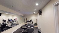 Located at 5461 Collins Mill Creek Drive in Milton, Florida, this single-floor fitness center is designed to provide a functional and well-equipped workout environment. The space features a selection of cardio machines, including ellipticals and a treadmill, arranged to maximize usability within the room. A large wall-mounted mirror along one side creates a sense of openness and allows users to monitor their form during exercise. Recessed lighting in the textured ceiling provides bright, even illumination throughout the space. A wall-mounted flat-screen television adds an entertainment element, making workouts more engaging. The flooring consists of durable, dark rubber matting suited for high-traffic fitness use. A wall-mounted fan ensures air circulation and comfort during activity. A small decorative plant near the equipment adds a touch of warmth to the otherwise utilitarian setting. The neutral-toned walls and clean layout contribute to a focused, distraction-free atmosphere, making this fitness center a practical and well-appointed space for residents or members seeking convenient access to exercise amenities.