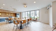 Located at 345 Lincoln Ave in Amherst, Massachusetts, this single-floor communal amenity space offers a thoughtfully designed environment suited for gathering, dining, and collaborative use. The open-plan layout accommodates multiple seating arrangements, including a large central communal table with modern metal-frame chairs, intimate booth-style seating upholstered in teal and pink, and smaller café-style tables positioned near expansive floor-to-ceiling windows. These oversized windows flood the interior with natural light and provide views of the surrounding outdoor courtyard and neighboring buildings.

The kitchenette area along one wall features warm wood-paneled cabinetry extending to the ceiling, complemented by slate-blue lower cabinets and a stone-effect countertop. Appliances include a stainless steel refrigerator, microwave, and sink, making the space functional for everyday use. Decorative gold dome wall sconces illuminate the kitchen zone, adding warmth against the natural wood backdrop.

A recessed lounge nook, framed by slatted wood ceiling details and a lush living green wall, creates an intimate retreat within the broader space. Light wood-look flooring runs throughout, unifying the various zones. The overall aesthetic balances contemporary design with natural materials, resulting in a welcoming and versatile communal environment.