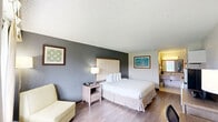 Located at 1400 W Tunnel Blvd in Houma, Louisiana, this efficiently designed guest room offers a comfortable and functional layout across a single floor. The bedroom area features hardwood-style flooring, a large upholstered headboard, and a generously sized bed dressed in crisp white linens. A wide window with decorative draping allows natural light to fill the space, while a cozy armchair and side lamp provide a relaxing corner for unwinding.

The room is well-equipped for productivity, featuring a dedicated work area with a spacious desk, ergonomic chair, and a media console with a flat-screen TV and chest of drawers. A compact kitchenette rounds out the living space, outfitted with white cabinetry, a mini-refrigerator, microwave, and countertop workspace — offering added convenience for extended stays.

The vanity area connects the main room to the bathrooms, featuring a broad granite-style countertop, an integrated sink, and open closet space with hanging storage and a luggage rack. A wall-mounted hair dryer adds a practical touch. The full bathroom includes tile flooring, neutral tile surround, a combined tub and shower with a curtain, and a toilet — designed with clean, utilitarian finishes. The overall ambiance is bright, neutral, and efficiently organized.
