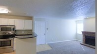 This single-floor space showcases an open-concept design that seamlessly connects the kitchen and living area. The kitchen features white cabinetry, a dark countertop bar with a built-in sink, offering a functional workspace that overlooks the main living space. The living area is finished with wall-to-wall carpeting, contributing to a warm and comfortable atmosphere. A tiled entryway is visible near the front door, providing a practical transition zone as you enter. The walls are painted in a neutral, light tone that enhances the sense of openness and brightness throughout the space. A textured popcorn ceiling spans the entire unit, adding a classic residential character. The paneled front door serves as the main entry point, accompanied by a thermostat panel mounted nearby on the wall, offering convenient climate control. Subtle natural light filters through the space, complementing the overall clean, neutral aesthetic. The layout is straightforward and functional, making efficient use of the available floor space while maintaining a cohesive flow between the kitchen and living zones.