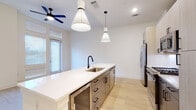 This contemporary single-floor residence showcases a clean, modern aesthetic throughout, with light wood flooring and crisp white walls that create a bright, cohesive atmosphere across all rooms. The open-concept kitchen serves as the centerpiece of the living area, anchored by a generously sized center island topped with white quartz countertops and fitted with an undermount stainless steel sink and a matte black faucet. Dual conical pendant lights hang above the island, complementing the recessed lighting overhead. The kitchen is well-appointed with two-tone cabinetry — light upper cabinets paired with darker lower drawers featuring matte black hardware — along with a full suite of stainless steel appliances, including a French door refrigerator with ice and water dispenser, a gas range, a built-in microwave, and a dishwasher. A white subway tile backsplash adds a refined touch. The living area benefits from tall windows that invite abundant natural light, enhanced by a ceiling fan for comfort. The bedroom is a calm, understated space with the same warm wood flooring and ample room. The bathroom is a standout, featuring large-format marble-look tile in the walk-in glass-enclosed shower alongside neutral tile flooring, all accented with matte black fixtures for a polished finish.