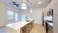 This contemporary single-floor residence showcases a clean, modern aesthetic throughout, with light wood flooring and crisp white walls that create a bright, cohesive atmosphere across all rooms. The open-concept kitchen serves as the centerpiece of the living area, anchored by a generously sized center island topped with white quartz countertops and fitted with an undermount stainless steel sink and a matte black faucet. Dual conical pendant lights hang above the island, complementing the recessed lighting overhead. The kitchen is well-appointed with two-tone cabinetry — light upper cabinets paired with darker lower drawers featuring matte black hardware — along with a full suite of stainless steel appliances, including a French door refrigerator with ice and water dispenser, a gas range, a built-in microwave, and a dishwasher. A white subway tile backsplash adds a refined touch. The living area benefits from tall windows that invite abundant natural light, enhanced by a ceiling fan for comfort. The bedroom is a calm, understated space with the same warm wood flooring and ample room. The bathroom is a standout, featuring large-format marble-look tile in the walk-in glass-enclosed shower alongside neutral tile flooring, all accented with matte black fixtures for a polished finish.