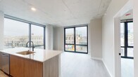 Located at 1110 W Wilson Ave in Chicago's Uptown neighborhood, this single-floor unit showcases a contemporary design aesthetic defined by exposed concrete ceilings, light hardwood-style flooring, and an open layout that maximizes both space and natural light. Large, floor-to-ceiling windows with black frames anchor the main living area, offering expansive views of the surrounding Uptown streetscape and flooding the interior with daylight.

The kitchen is a standout feature, outfitted with wood-toned cabinetry, a white quartz waterfall island with an integrated sink and matte black faucet, subway tile backsplash, and stainless steel appliances including a gas range, microwave, and refrigerator. The kitchen flows seamlessly into the open living area, making it well-suited for everyday living and entertaining.

A hallway leads to the private areas of the unit, including a bedroom with a built-in sliding-door closet featuring integrated shelving and hanging rods. Two bathrooms are finished with large-format tile flooring, wood-grain vanities, white countertops, and chrome hardware. White walls throughout contribute to a clean, minimalist ambiance that complements the unit's modern industrial character.