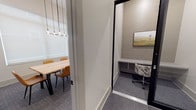 This single-floor office suite is thoughtfully designed to accommodate a variety of work styles, combining open collaboration with private, focused workspaces. The centerpiece of the suite is a well-appointed conference room featuring soaring ceilings, a substantial natural wood table surrounded by tan leather chairs, and an eye-catching cluster pendant light fixture that adds a contemporary aesthetic touch. Large windows fitted with roller shades allow abundant diffused natural light to fill the room, creating a bright and welcoming atmosphere. Landscape artwork adorns the walls, contributing to a calm, professional ambiance throughout. Adjacent to the conference room, a corridor leads to a series of compact, glass-enclosed private focus pods framed in sleek black metal. Each pod is equipped with a built-in desk surface and an ergonomic office chair, providing a quiet, distraction-free environment ideal for individual tasks or private calls. The glass walls maintain visual openness while offering acoustic separation. Patterned blue-grey commercial carpet flows throughout the suite, tying the spaces together with a cohesive, polished look. A built-in countertop workspace along one wall adds additional utility to the shared corridor area, making this suite a versatile and functional professional environment.