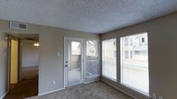 This single-floor space presents a straightforward and functional layout designed for comfortable everyday living. The main living area is defined by neutral-toned walls paired with wall-to-wall carpeting underfoot, creating a cohesive and understated aesthetic throughout. A textured popcorn ceiling adds a classic finish overhead.

Natural light plays a prominent role in this space, with a large picture window along one wall allowing generous daylight to fill the room. Fitted with horizontal blinds, the window offers flexible light control while maintaining views of the surrounding exterior. An exterior door, also featuring a full-length glass panel and matching blinds, provides direct access to the outdoors and further enhances the sense of connection to the outside environment. Together, the window and glass door contribute to a bright and open atmosphere.

The room's layout is open and unobstructed, offering flexibility for a variety of furniture arrangements. The neutral color palette of the walls, flooring, and trim creates a versatile backdrop suitable for a range of interior styles. Overall, the space presents a clean, practical environment with ample natural light and straightforward design elements.