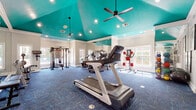 This fitness center makes a bold architectural statement with its vaulted teal ceiling, which soars overhead and is accented by recessed lighting and ceiling fans that keep the space bright and comfortable. Crown molding lines the transition between the walls and ceiling, lending a polished, upscale feel to the workout environment.

The main exercise area is generously proportioned, offering ample room for a comprehensive array of equipment. Cardio machines, including multiple treadmills, stationary bikes, and an elliptical, are arranged throughout the space, while a multi-function cable machine and a full dumbbell rack support strength training. Floor-to-ceiling mirrors line the walls, visually expanding the room and providing exercisers with proper form feedback. Patterned blue carpet tiles cover the floors, adding color and a degree of cushioning underfoot.

Large windows along the perimeter flood the space with natural light and offer outdoor views, creating an open and energizing atmosphere. Medicine balls, stability equipment, and wall-mounted fitness reference charts round out the functional amenities.

Adjacent to the main workout floor, a dedicated steam room finished entirely in elegant marble-patterned tile features a built-in bench and mosaic tile flooring, providing a spa-like recovery experience that elevates the overall fitness facility.