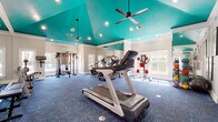 This fitness center makes a bold architectural statement with its vaulted teal ceiling, which soars overhead and is accented by recessed lighting and ceiling fans that keep the space bright and comfortable. Crown molding lines the transition between the walls and ceiling, lending a polished, upscale feel to the workout environment.

The main exercise area is generously proportioned, offering ample room for a comprehensive array of equipment. Cardio machines, including multiple treadmills, stationary bikes, and an elliptical, are arranged throughout the space, while a multi-function cable machine and a full dumbbell rack support strength training. Floor-to-ceiling mirrors line the walls, visually expanding the room and providing exercisers with proper form feedback. Patterned blue carpet tiles cover the floors, adding color and a degree of cushioning underfoot.

Large windows along the perimeter flood the space with natural light and offer outdoor views, creating an open and energizing atmosphere. Medicine balls, stability equipment, and wall-mounted fitness reference charts round out the functional amenities.

Adjacent to the main workout floor, a dedicated steam room finished entirely in elegant marble-patterned tile features a built-in bench and mosaic tile flooring, providing a spa-like recovery experience that elevates the overall fitness facility.