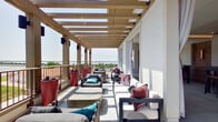 This thoughtfully designed single-floor space blends modern aesthetics with coastal influences throughout its distinct areas. The expansive covered terrace is a standout feature, stretching along the building's exterior beneath a slatted wood pergola that filters natural sunlight. Furnished with plush, cushioned seating in neutral and jewel tones, the terrace opens to panoramic waterfront views, creating an inviting outdoor retreat ideal for relaxing or entertaining.

Inside, the kitchen is bold and visually striking, anchored by cobalt blue cabinetry paired with a light quartz countertop island. Patterned geometric floor and wall tiles add graphic energy, while a series of sleek pendant lights illuminate the space from above. Wicker bar stools line the island, and large-scale coastal artwork adorns the walls, reinforcing the waterfront theme.

The living area offers a refined contrast, featuring light hardwood flooring, soaring ceilings, and a dramatic floor-to-ceiling dark metal fireplace surround. Large black-framed glass doors and windows flood the room with natural light and provide direct terrace access.

A separate lounge area incorporates high-top bar tables, stone accent walls, warm wood paneling, and water views visible through generous windows, completing this well-appointed and cohesive space.