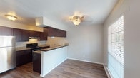 This single-story, two-bedroom, one-bathroom unit offers a practical and well-appointed layout across a comfortable footprint. The open-concept living and dining area is anchored by wood-style plank flooring that runs throughout the main living spaces, lending warmth and visual continuity. Ceiling fans are present in the living area and bedrooms, providing both function and a finished look. Natural light enters through multiple windows fitted with horizontal blinds, brightening the neutral-toned walls.

The kitchen is efficiently designed in a galley configuration, featuring rich dark-wood cabinetry that extends to the upper walls for ample storage. Granite-style countertops line both sides of the kitchen, complementing the black appliances including an electric range. A peninsula with a raised bar-height counter separates the kitchen from the dining and living area, creating an open yet defined transition between spaces.

Both bedrooms are comfortably sized, carpeted for comfort, and include ceiling fans and windows that allow natural light. The shared bathroom includes a dark-wood vanity with a granite-style countertop, a single-basin sink, and a full tub-shower combination, all set on tile flooring. The overall design is cohesive, clean, and functional.