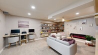 Located at 8040 Rowland Avenue in Philadelphia, PA, this single-floor library and lounge space is a well-appointed retreat designed for reading, relaxation, and productivity. The room is finished with soft neutral tones throughout, including light gray walls, white trim, and plush wall-to-wall carpeting that contributes to a warm and inviting atmosphere.

The defining feature of the space is its extensive built-in wooden shelving that lines multiple walls from floor to near-ceiling, providing substantial storage for a large personal book collection. These open shelves wrap around two primary walls, creating an immersive library experience. Centrally positioned against one wall is a sleek, horizontal electric fireplace with a vivid color-changing display, flanked by framed artwork and decorative accents that add personality to the space.

The lounge area is furnished with a modern white sofa and matching armchairs accented with mauve throw pillows, arranged around a minimalist wood coffee table — ideal for comfortable reading or conversation. A stylish ceiling fan with gold-toned blades provides both function and aesthetic detail overhead, complemented by recessed lighting throughout.

A dedicated workstation with a floating wooden desk and computer setup occupies one corner, making the space equally suited for focused work. A glass door element is also visible, adding a touch of architectural refinement. Overall, the space balances functional design with a cozy, curated ambiance.