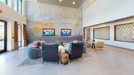Located in Richmond, Virginia, this single-floor community clubhouse delivers a refined blend of contemporary design and thoughtful craftsmanship. The centerpiece of the space is a soaring double-height lounge area bathed in natural light through floor-to-ceiling windows, offering direct views of the outdoor pool area. A sweeping large-scale abstract mural anchors one wall, flanked by dual wall-mounted televisions, while plush curved sofas and sculptural lounge chairs create an inviting social atmosphere. Distinctive architectural accents are found throughout, including decorative latticed wall panels framed in warm wood set against geometric patterned tile flooring, adding artful texture to transitional areas. The kitchen features rich walnut cabinetry, dark stone countertops, and stainless steel appliances, complemented by a sculptural 3D white tile backsplash and sleek pendant lighting. A marble-clad feature wall with gold veining adds a luxurious focal point near the bar seating area, where a polished stone countertop and leather chairs invite casual gatherings. Recessed lighting and pendant fixtures provide warm illumination throughout, while the cohesive palette of natural stone, warm wood tones, and neutral walls creates a sophisticated and welcoming environment.
