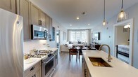 Located at The Vanderbilt in Westbury, New York, this single-floor residence offers a thoughtfully designed open-concept layout that seamlessly connects the kitchen, dining, and living areas into one cohesive living space. The kitchen is a standout feature, appointed with taupe cabinetry, white subway tile backsplash, quartz countertops, a gold-toned undermount sink, and stainless steel appliances including a gas range and built-in microwave. Pendant lighting adds a refined touch above the counters. The open flow leads directly into a dedicated dining area, which transitions naturally into a comfortable living room bathed in natural light from large windows dressed with bright curtains. The living space is finished with light hardwood-style flooring that runs throughout, contributing to a warm and airy ambiance. Recessed lighting overhead ensures the space remains well-lit at all hours. The neutral color palette — featuring soft whites, warm grays, and natural wood tones — creates a clean, modern aesthetic throughout. The Vanderbilt offers a sophisticated residential experience in the heart of Westbury, combining contemporary design with functional, everyday comfort.