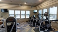This fitness center is thoughtfully designed to support an active lifestyle, offering a bright and inviting atmosphere for residents or members. The room is lined with large windows along one wall, flooding the space with natural light and providing open views of the surrounding landscape, creating an airy feel during workouts. Two ceiling fans are suspended overhead, ensuring comfortable airflow throughout the room.

The flooring features a distinctive herringbone-patterned carpet in a mix of neutral and muted tones, adding a stylish yet practical foundation to the space. A wall-mounted flat-screen television is positioned for easy viewing from most cardio equipment, enhancing the workout experience.

The fitness center is equipped with a variety of modern cardio machines, including ellipticals, stair climbers, and additional exercise equipment, all arranged to maximize usable floor space while maintaining an open, uncluttered layout. Recessed lighting supplements the natural light, ensuring the space remains well-lit at all hours. The walls feature a neutral color palette with subtle horizontal banding, contributing to a clean and polished aesthetic throughout.