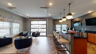 This thoughtfully designed single-floor amenity space blends modern aesthetics with versatile functionality. The open-plan layout is anchored by a striking herringbone-patterned hardwood floor that flows throughout, tying together the various zones with visual cohesion.

At the heart of the space is a long bar counter with high-top seating, illuminated by a row of elegant brass-and-glass pendant lights suspended from the ceiling. Adjacent to it, a full kitchenette features bold orange cabinetry, black granite countertops, stainless steel appliances, and a dramatic textured geometric accent wall with dual wall-mounted flat-screen televisions — ideal for entertaining or casual gatherings.

A comfortable lounge area is furnished with plush navy velvet accent chairs, ottomans, and a layered rug, positioned to take advantage of the abundant natural light streaming through large windows dressed with decorative roman shades. A defining architectural feature is the full-height glass panel garage-style door, which opens to an outdoor balcony and floods the interior with light while offering seamless indoor-outdoor connectivity.

A private meeting nook, enclosed with its own entry door, accommodates a round wood table surrounded by leather chairs and its own wall-mounted display screen — perfect for small group discussions or focused work sessions. Recessed ceiling lighting throughout ensures the space is well-lit at all hours.