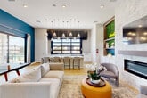 Located at 20807 W Indian School Rd in Buckeye, Arizona, this modern community clubhouse is a well-appointed single-floor gathering space designed for socializing and relaxation. The open-concept layout seamlessly blends several functional zones within a thoughtfully curated interior.

The living area anchors the space with a plush sectional sofa, accent chairs, and a bold yellow ottoman, all centered on a patterned area rug over warm-toned hardwood flooring. A striking marble-clad feature wall houses a sleek linear fireplace beneath a wall-mounted flat-screen television, flanked by a lush vertical living wall with illuminated shelving — a standout design element that adds organic texture and color.

The kitchen bar area features a long white island with seating for multiple guests, pendant lighting, and a large industrial-style window that floods the space with natural light and frames outdoor views of the pool area. White cabinetry and clean countertops complete the crisp, contemporary aesthetic.

Bold teal accent walls punctuate the otherwise neutral palette, creating a vibrant energy throughout. A dining nook with curated wall art, a ping pong table, and sliding glass doors opening to the pool deck round out the space, making it a versatile hub for both leisure and community connection.