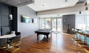 Located at 3635 NE 1st Ave in Miami's vibrant Midtown neighborhood, this contemporary amenity lounge offers a well-appointed space designed for entertainment and socializing. Rich hardwood flooring runs throughout, anchoring the room's modern aesthetic and complementing the cool-toned gray walls.

At the heart of the space sits a full-sized pool table, accompanied by a wall-mounted cue rack and flanked by wall-mounted flat-screen TVs, creating an ideal setting for leisure and recreation. A dedicated lounge area with seating provides a comfortable retreat within the same open layout.

The bar area is a standout feature, showcasing a sleek concrete countertop and base clad in bold, vivid blue subway tiles, adding a striking pop of color to the industrial-modern palette. Gold-accented bar stools line the counter, while cylindrical pendant lights hang overhead. Behind the bar, a dramatic large-scale artistic wall mural of a geometric portrait serves as a commanding focal point, adding a gallery-like quality to the space.

Floor-to-ceiling sheer-draped glass doors and windows flood the interior with natural light and offer views of the surrounding Midtown Miami streetscape. The recessed lighting and layered ceiling details further elevate the refined, urban character of this thoughtfully designed communal space.